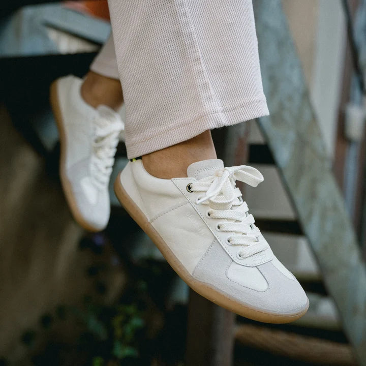 Close-up of a person wearing BluSun BLSN-200 (Men’s) white organic cotton sneakers with gum soles and light-colored pants, sitting on a metal staircase with legs dangling.