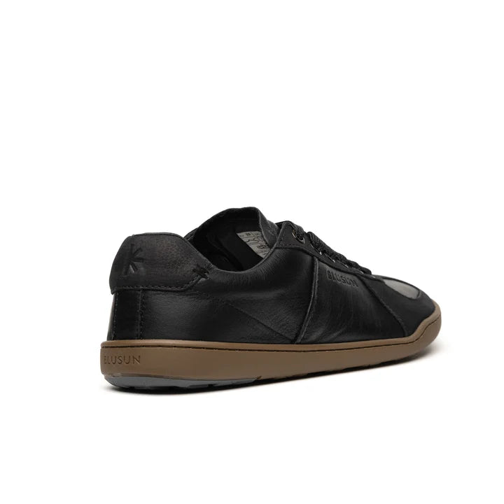 A black leather BLSN-200 men’s sneaker by BluSun, featuring a brown rubber sole, is shown from the back left angle. The heel showcases a subtle embossed logo, while “BluSun” appears on the edge of the sole. This leather shoe is lined with organic cotton for enhanced comfort and breathability.
