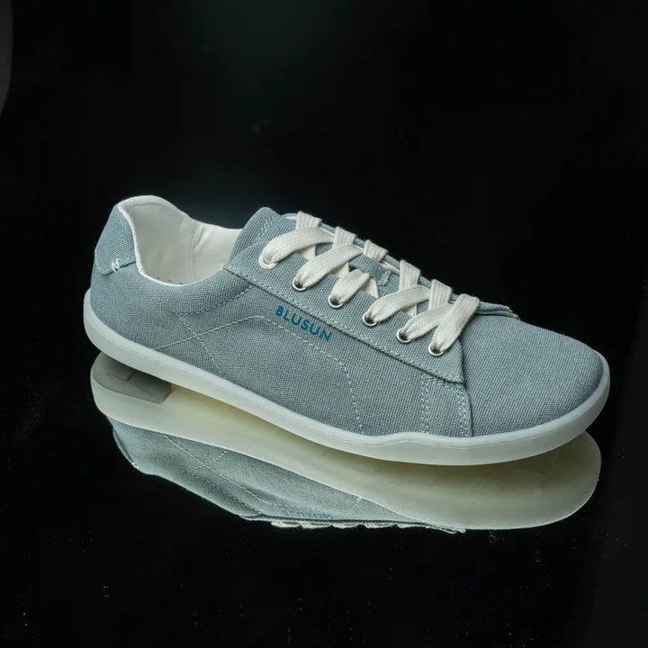 Introducing the BluSun BLSN-103 (Women’s)—a light gray canvas sneaker crafted from organic cotton for barefoot comfort and sustainability. This shoe features white laces, a white sole, and the brand name BLUSUN in blue on the side. Presented on a glossy black surface with its reflection visible.