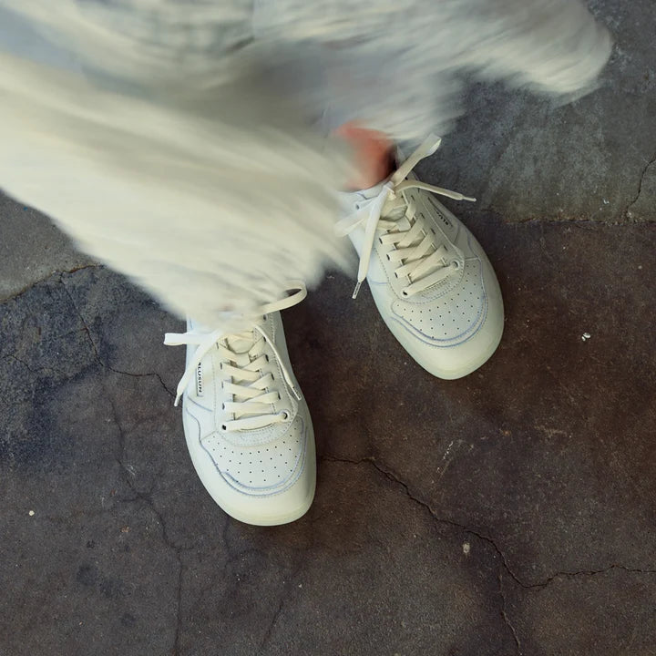 A person wearing BluSun BLSN-600+ women’s smooth leather white sneakers with a wide toe box stands on a cracked concrete surface, partially covered by a flowing, light fabric or dress.