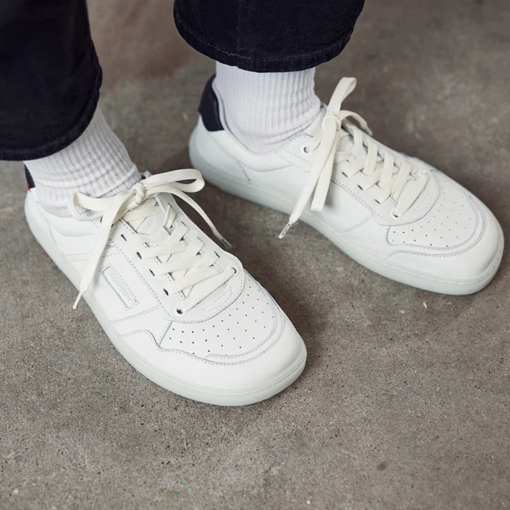 A person wearing BluSun BLSN-600+ men’s barefoot sneakers with a wide toe box and white socks stands on a gray concrete floor. Only the lower legs and feet are visible, with dark pants emphasizing the natural comfort design of the BluSun footwear.