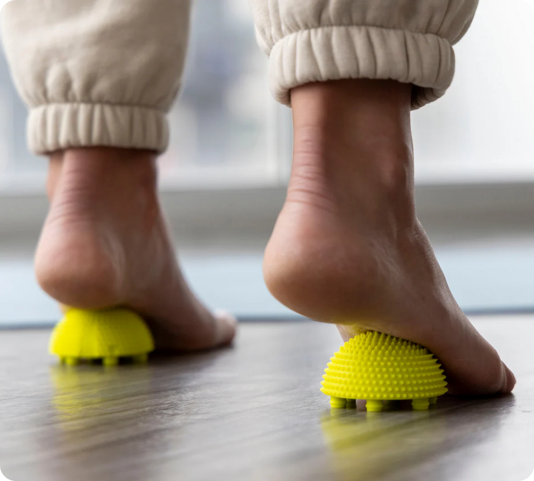 A person in beige pants presses bare feet onto yellow NABOSO Neuro Balls on a wooden floor, likely performing a calming foot massage or exercise.
