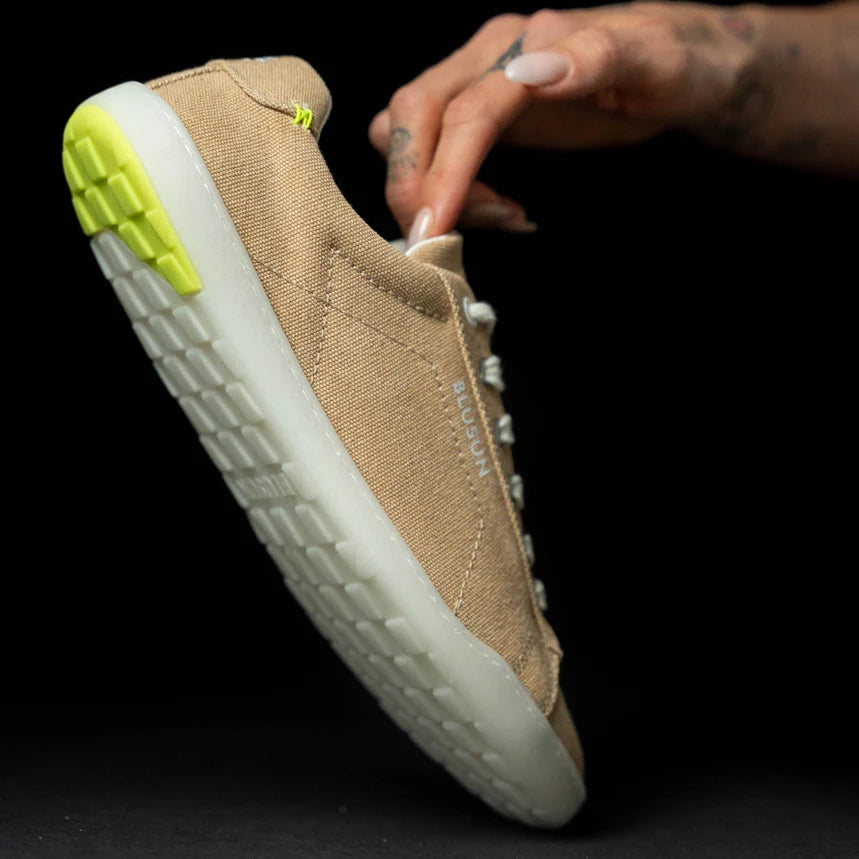 A hand holds the BluSun BLSN-103 Men’s sneaker—a beige, eco-friendly vegan shoe with a white sole and yellow heel accent—displayed against a black background and designed for both style and sustainability.