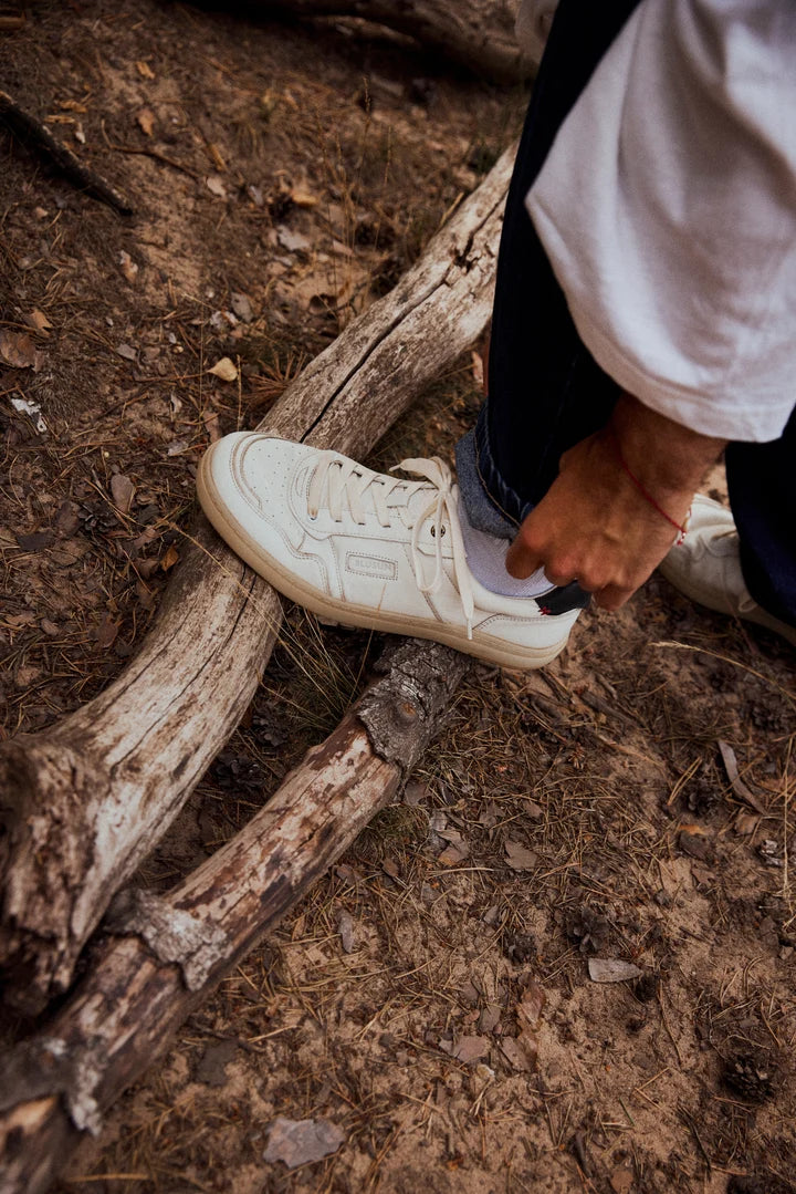 Wearing BluSun BLSN-600+ men’s barefoot sneakers with a wide toe box and blue jeans, a person steps on a fallen tree branch in a wooded area, enjoying the natural comfort these shoes offer.