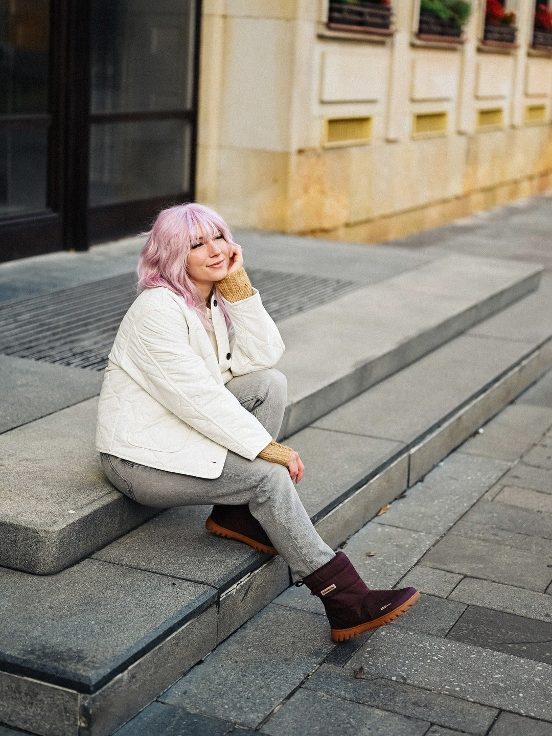 A person with pastel pink hair sits on outdoor steps, smiling with eyes closed. They're wearing Barebarics Polarstride (Women's) brown winter boots with a waterproof membrane, plus a white quilted jacket, gray pants, and a tan sweater.