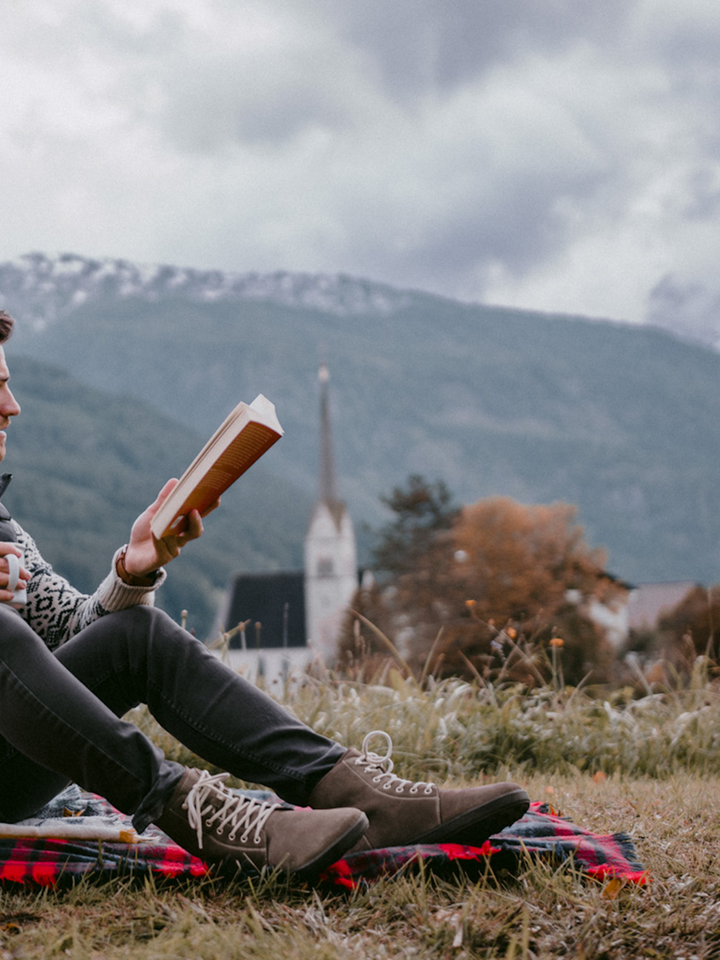 Relaxing on a blanket outdoors, someone reads a book in BeLenka Winter 3.0 barefoot shoes with Merino wool lining, surrounded by tranquil mountains, a church, autumn trees, and lush grass under cloudy skies.